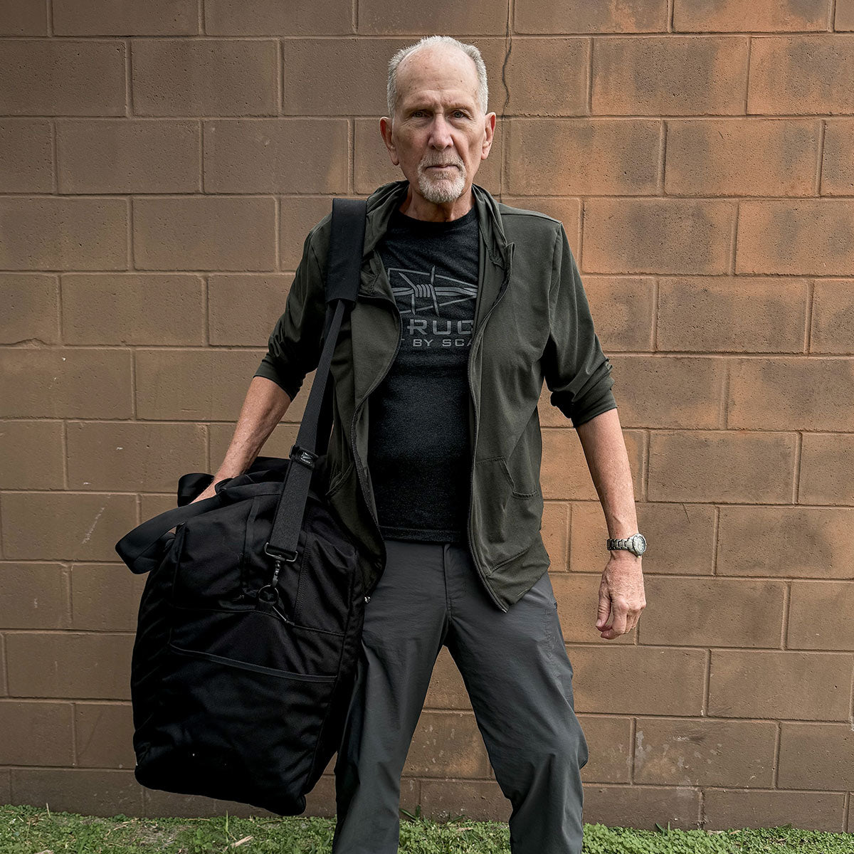 A man with short gray hair stands against a brick wall, holding a large Kit Bag - 84L from GORUCK. He wears a dark jacket over a T-shirt and pants, with a watch on his left wrist. Grass covers the ground beneath him.