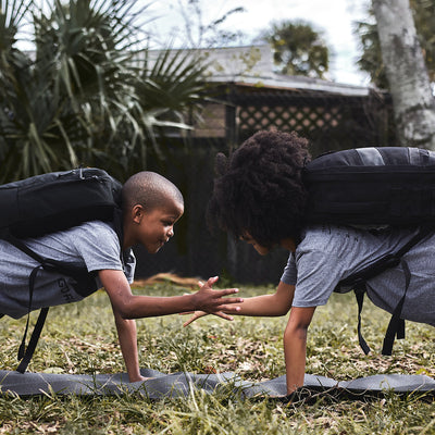 In a grassy area surrounded by palm trees, two children participating in the GORUCK Elementary School Ruck Training Program are in a garden plank position, high-fiving each other with their free hands as they master functional movements while wearing backpacks.