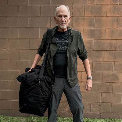 An elderly man in a dark jacket stands against a brick wall, clutching a Kit Bag (Includes Shoulder Strap).