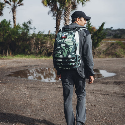 A person dressed in a gray outfit and black cap strolls along a gravel path beside a puddle, carrying the GORUCK GR1 USA x Gear Patrol rucksack in variegated elephant ear camo. Palm trees and greenery create a lush backdrop.