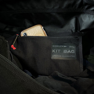 An open GORUCK Kit Bag - 84L (Includes Shoulder Strap) in black showcases a smartphone in a beige case snugly placed in the side pocket, with its capacity label clearly visible. Its design highlights durability and organization, catering to the requirements of challenging deployments.