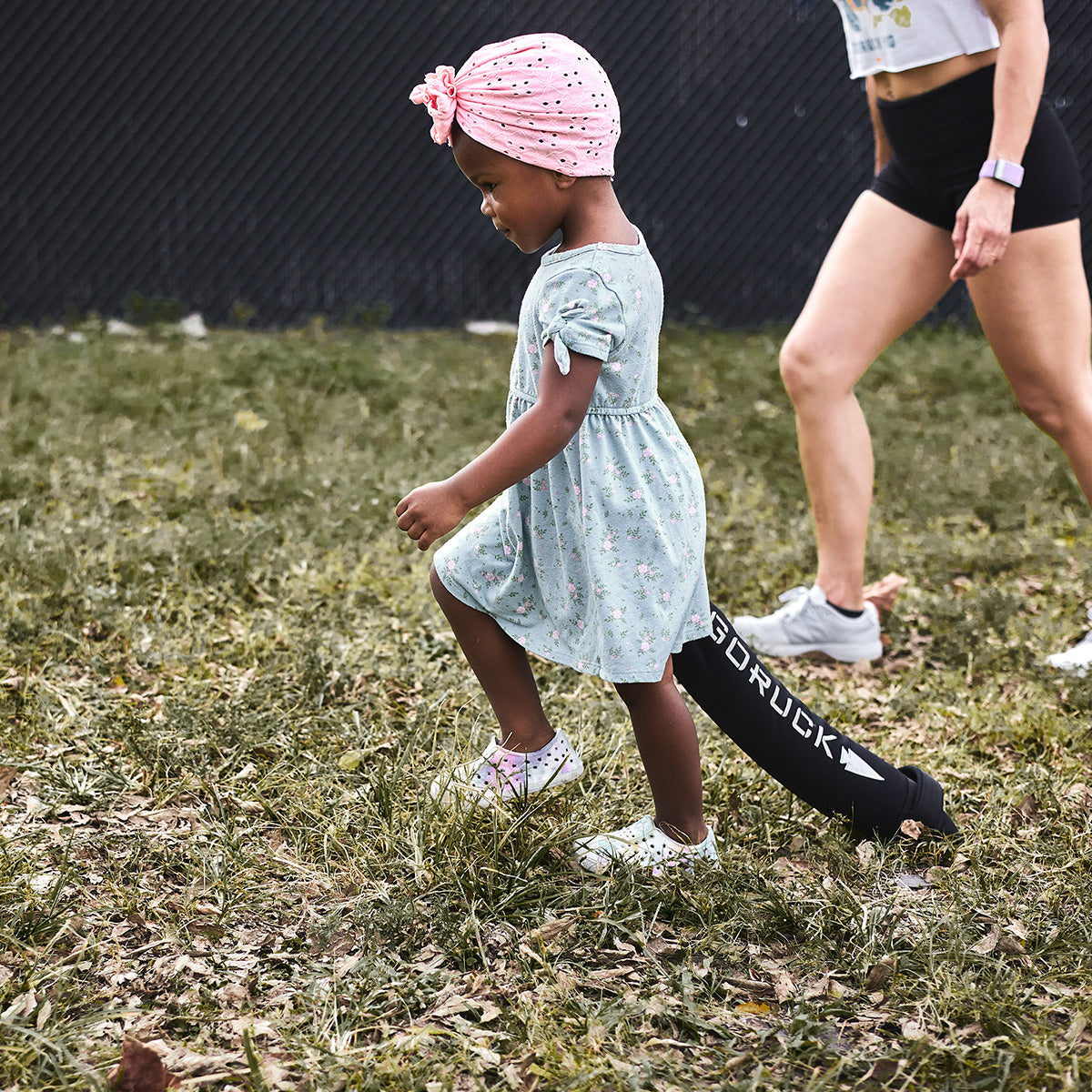 A young child in a pink headscarf and floral dress navigates over a black obstacle branded with GORUCK, reflecting the core ideas of functional movements from the Elementary School Ruck Training Program. Nearby, an adult dressed in athletic wear strolls on the grassy ground.