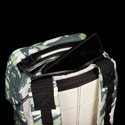 The GR1 USA x Gear Patrol by GORUCK, a limited-edition camouflage backpack inspired by variegated elephant ear camo, is slightly unzipped to reveal a black laptop inside. It features padded straps and a checkered interior, contrasted against a solid black background.