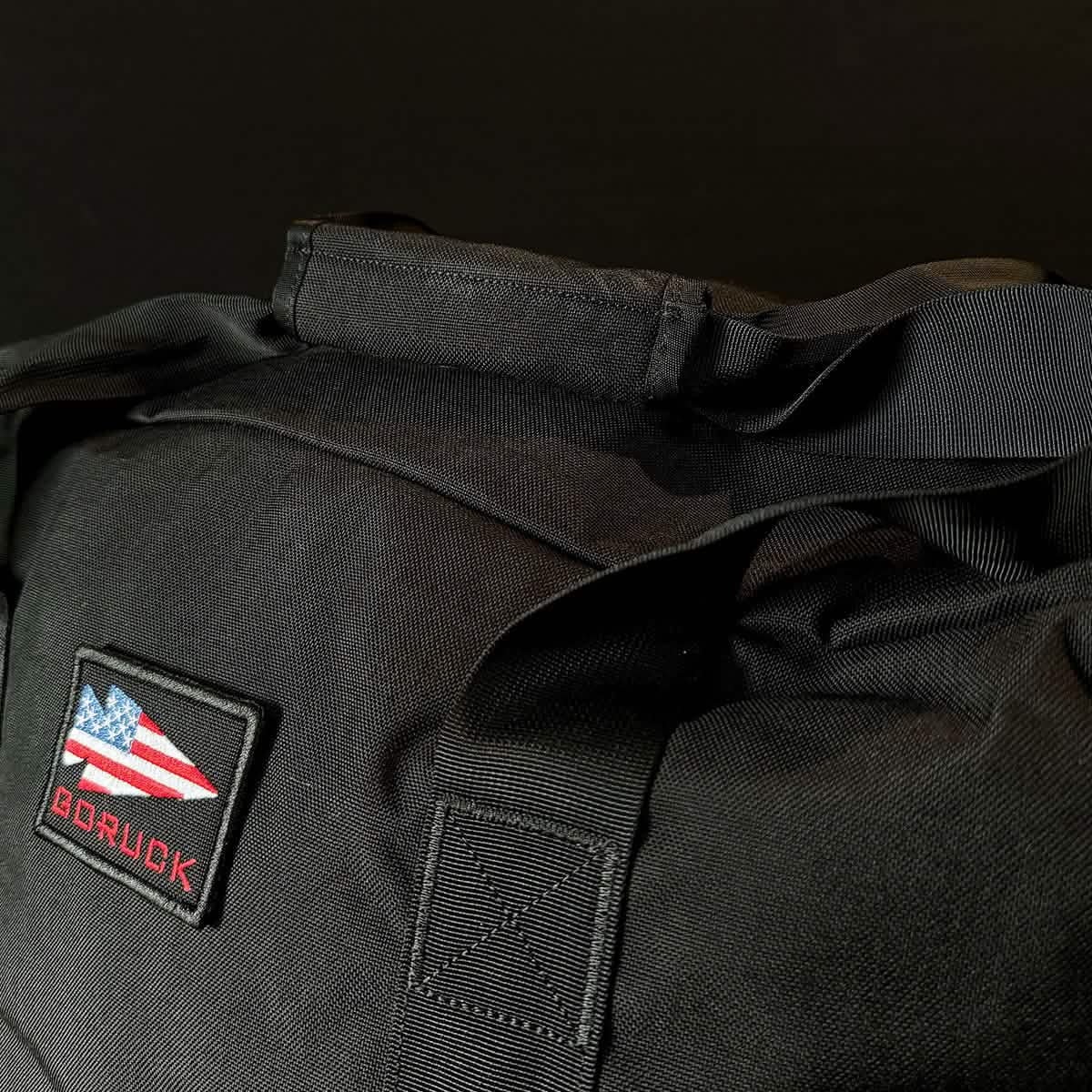 A close-up of a black Kit Bag (Includes Shoulder Strap) featuring a stylized American flag patch and the word "GORUCK," inspired by the rugged durability of Special Forces gear.