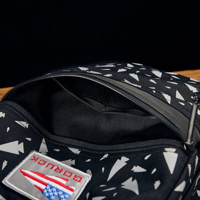 Close-up of the KR1 - Spearhead Splatter kids rucksack in black, featuring a triangular pattern. The open zipper offers a peek inside. A patch displaying an American flag design and the GORUCK brand name is sewn onto the fabric. Ideal for carrying school supplies, the backdrop enhances its sleek appearance.