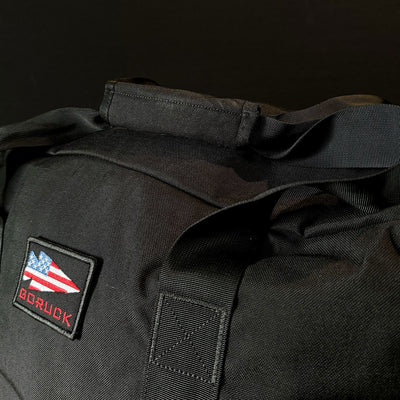 Close-up of the GORUCK Kit Bag - 84L, featuring a black design against a dark background and reminiscent of an Aviator’s Kit Bag. The bag displays a sewn patch with an American flag and the brand name GORUCK in red text.