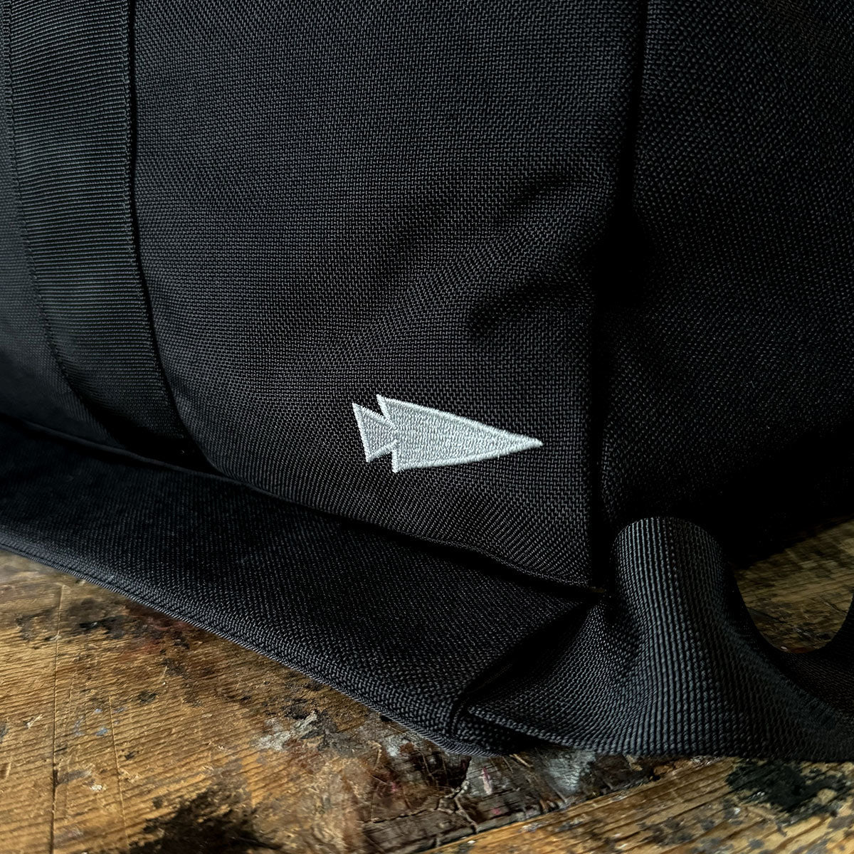 A close-up of a black fabric, reminiscent of the GORUCK Kit Bag - 84L (Includes Shoulder Strap), showcases a small embroidered white arrow symbol. The fabric is slightly folded at the bottom and rests gracefully on a worn wooden surface.