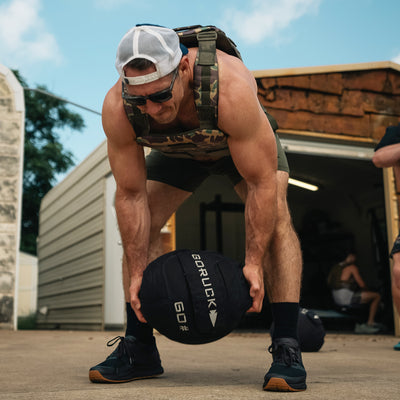 A muscular man in a camouflage vest and cap gets ready to lift a 60 lb GORUCK Sand Medicine Ball, enhancing his core stability. He stands before a garage with doors partially open under a clear sky, indicating that this outdoor area serves as his home gym for intense workouts.