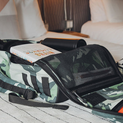 A limited-edition GR1 USA x Gear Patrol rucksack from GORUCK in variegated elephant ear camo lies open on a bed, revealing a book titled "Your Brain" and a black water bottle inside. Neutral-toned bedding is visible in the background.