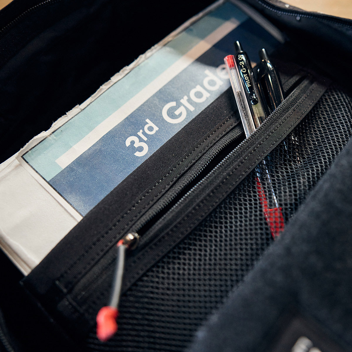 A GORUCK KR1 - Spearhead Splatter kids rucksack reveals a folder labeled 3rd Grade and several pens neatly tucked in a mesh pocket, brimming with essential school supplies.