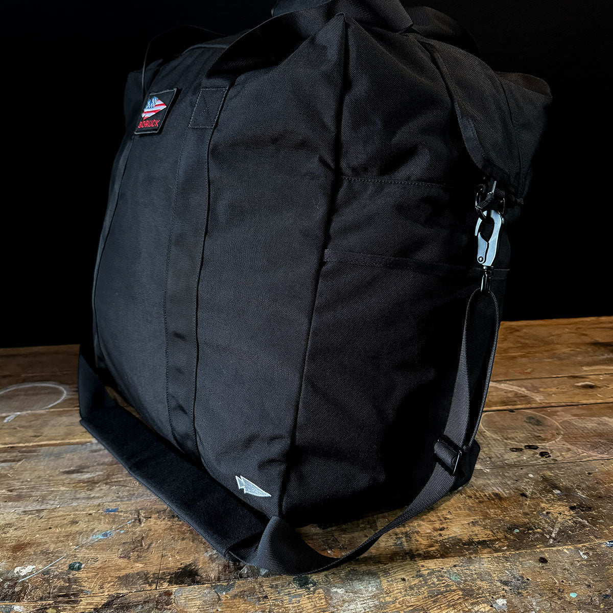 A black GORUCK Kit Bag - 84L, reminiscent of an Aviator’s Kit Bag, features a shoulder strap and metal clasp resting on a wooden surface. The small logo patch near the top adds subtle detail. A dark background accentuates its Special Forces Flight Bag-like design and texture.