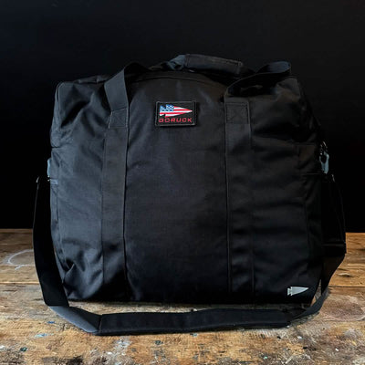 A black Kit Bag (Includes Shoulder Strap) by GORUCK, with an American flag patch, rests on a wooden surface against a dark background.