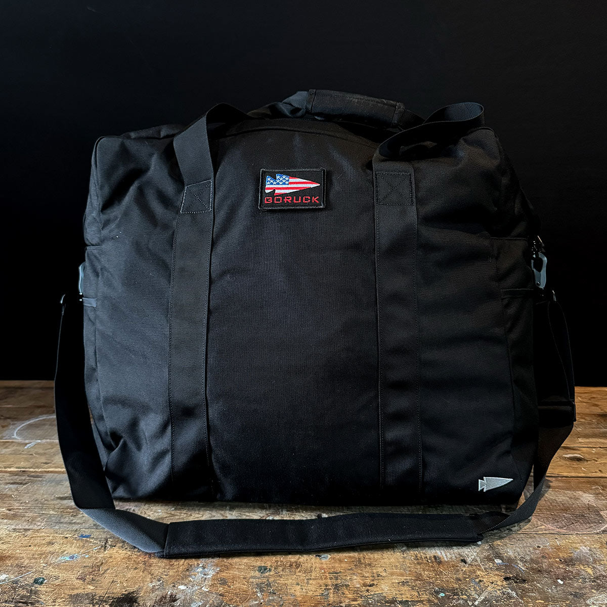 A black GORUCK Kit Bag - 84L, complete with a shoulder strap, is displayed against a dark background, evoking the design of an Aviator’s Kit Bag. The bag showcases an American flag patch above the GORUCK brand name. It is equipped with sturdy handles and rests on a wooden surface.