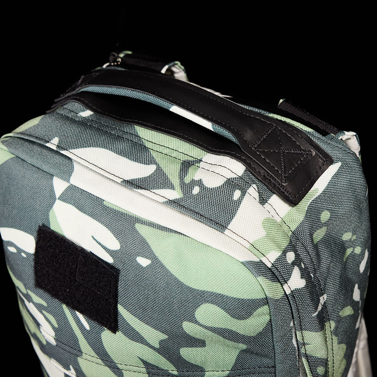 The GR1 USA x Gear Patrol rucksack from GORUCK showcases a unique variegated elephant ear camo pattern in green, white, and black. It features a black leather handle and a front patch area, all against an elegant solid black backdrop.