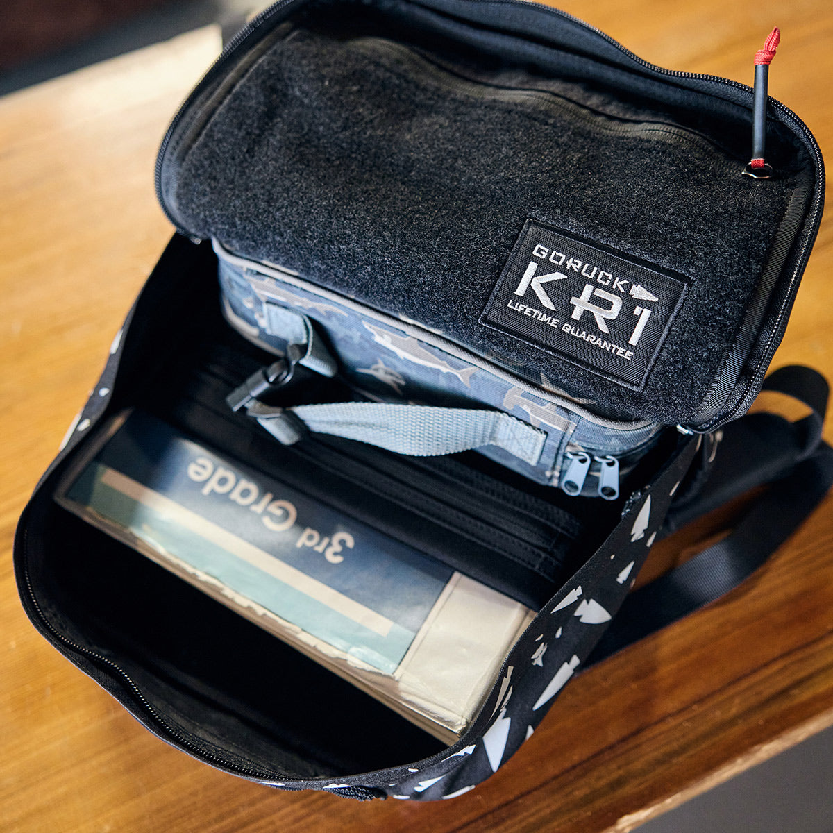 An open KR1 - Spearhead Splatter rucksack for kids rests on a wooden table, displaying a GORUCK kit with a Lifetime Guarantee label and a binder labeled 3rd Grade, ideal for organizing school supplies.