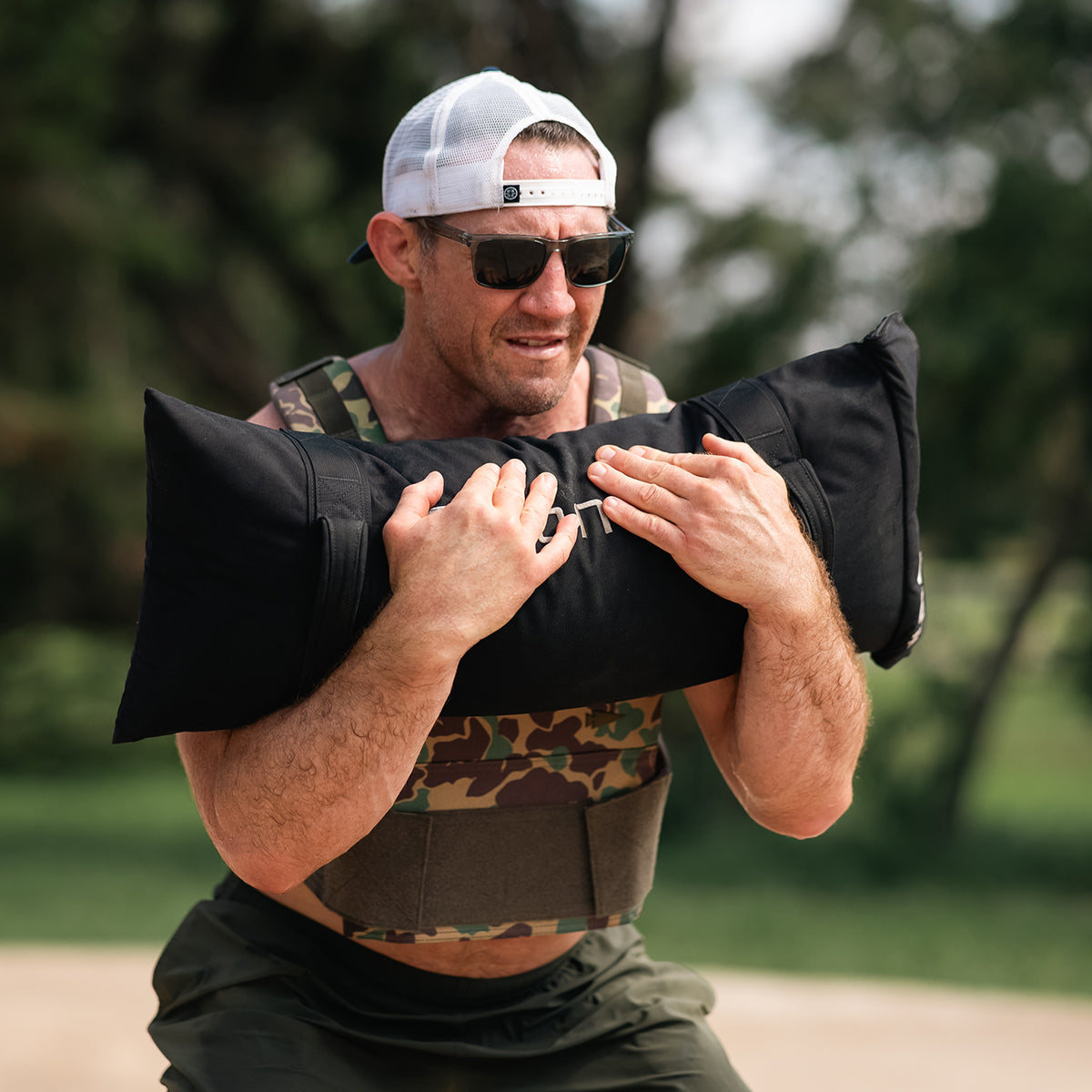 A man in a camouflage vest, sunglasses, and a white cap is outdoors, performing a squat with Simple Training Sandbags from GORUCK, which feature padded handles. Trees and greenery are visible in the background.