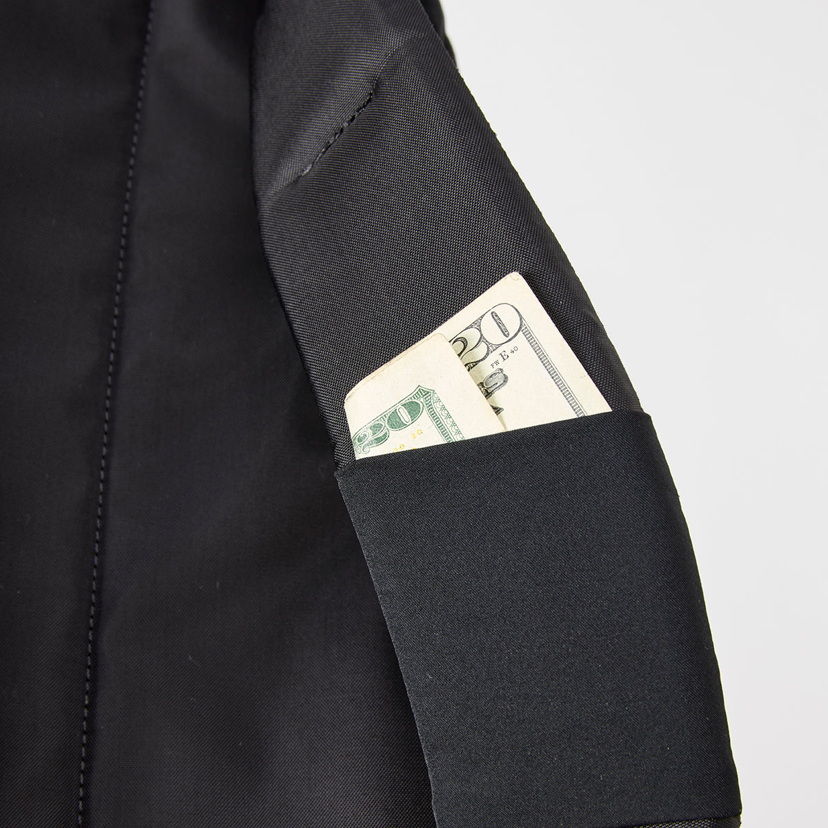 Twenty and ten-dollar bills peek from the black pocket of a Bullet Ruck - Ripstop ROBIC® backpack by Michael Easter, made from durable 420D ROBIC® Ripstop Nylon, against a plain backdrop.