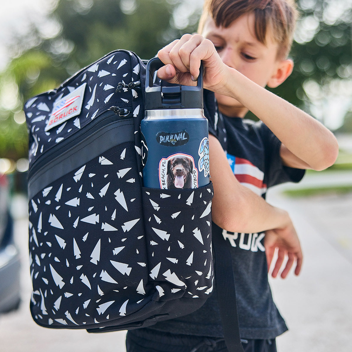 A young boy slides a water bottle, decorated with a chimpanzee sticker, into the side pocket of his GORUCK KR1 - Spearhead Splatter kids rucksack with its black and white geometric pattern. Wearing a dark t-shirt, he stands outside amidst the trees, prepared for both adventure and school supplies.