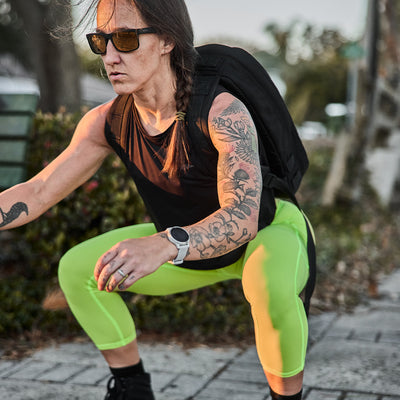 A person in sunglasses squats outdoors, wearing a black tank, neon green Women's Cropped Training Leggings Pocket made by GORUCK with ToughFlex Fabric, and a backpack.