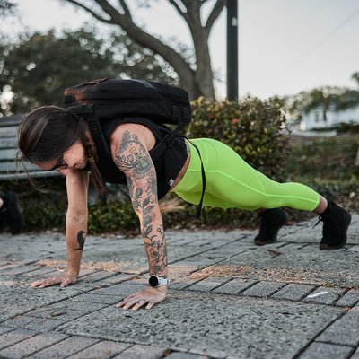 A tattooed individual does push-ups outside, wearing a backpack and vibrant green high-waisted Women's Cropped Training Leggings with Pocket made from GORUCK's ToughFlex Fabric.