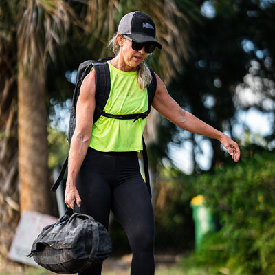 A woman dressed in a Women’s USA Performance Tank made from GORUCK's ToughMesh™ fabric in neon green, coupled with black leggings, a cap, and sunglasses, walks outdoors carrying a black duffel bag. As she wanders through the lush greenery and trees with her backpack on, her lightweight training shirt dries incredibly fast.