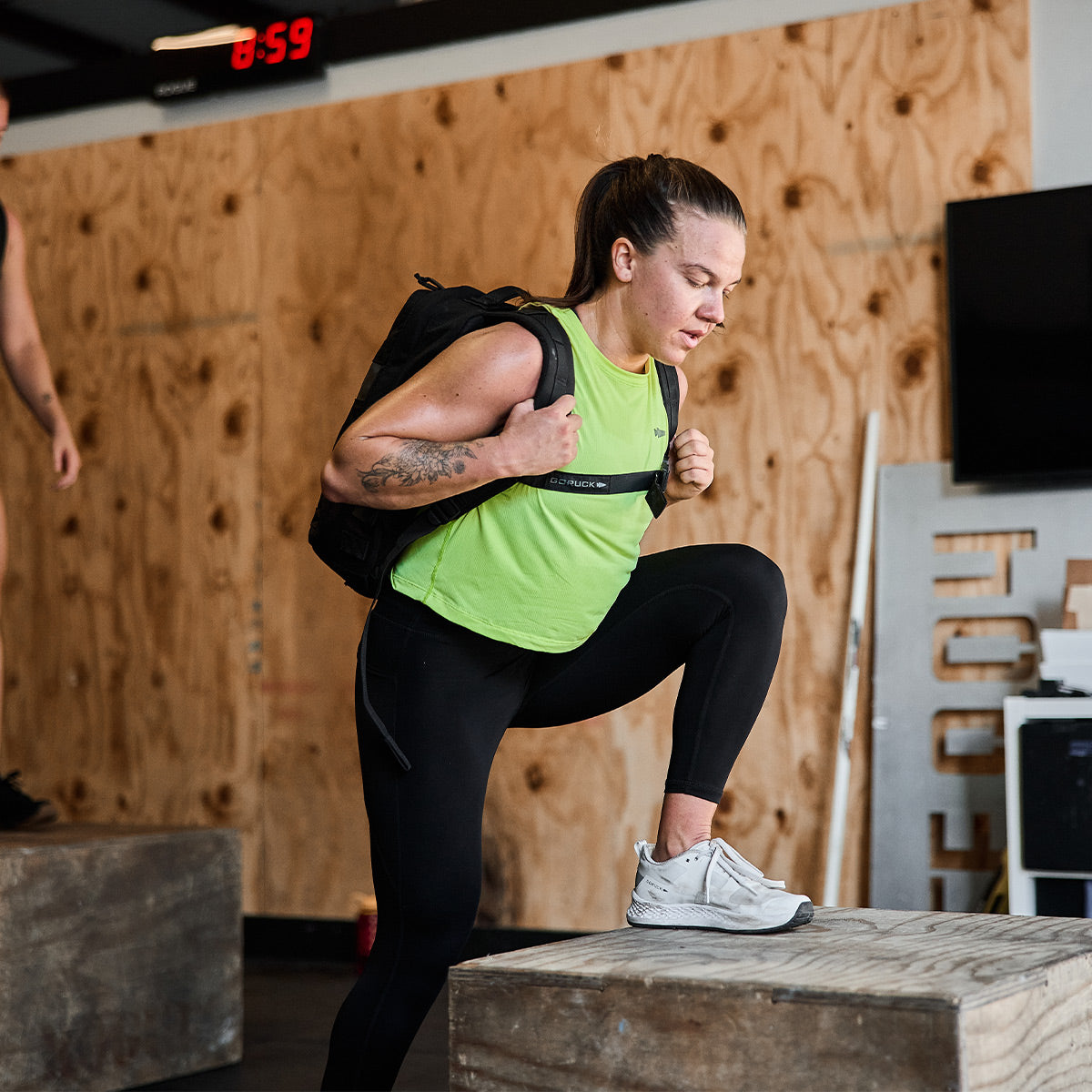 A woman, dressed in a green Women's USA Performance Tank - ToughMesh by GORUCK and black leggings, wearing a featherweight training shirt, performs step-ups on a wooden box with a backpack in a gym with plywood walls and a digital clock in the background.