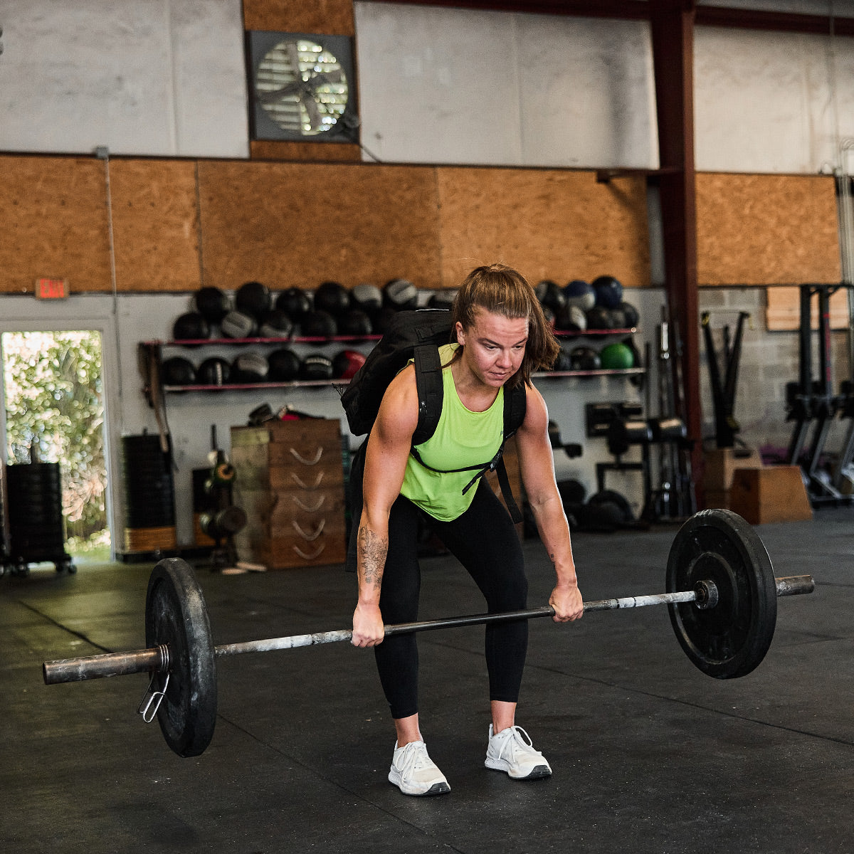 A person in a green Women’s USA Performance Tank - ToughMesh from GORUCK and black leggings lifts a barbell in the gym. The background features shelves with kettlebells and fitness equipment, while the space has a high ceiling and a fan.