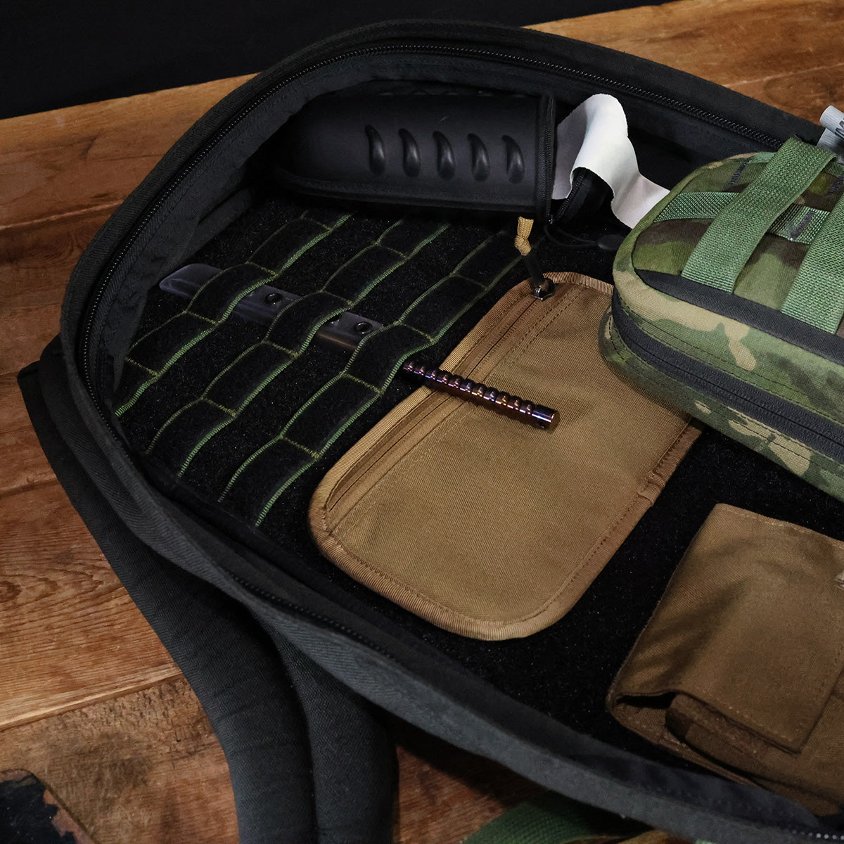 The Admin Panels by GORUCK lay open on the wooden floor, showcasing perfectly organized compartments ideal for EDC and rucking. Inside, there is a green case with a camouflage pattern, a brown pouch, a flashlight, and a tactical knife handle. The interior is expertly designed for efficiently carrying gear or tools.