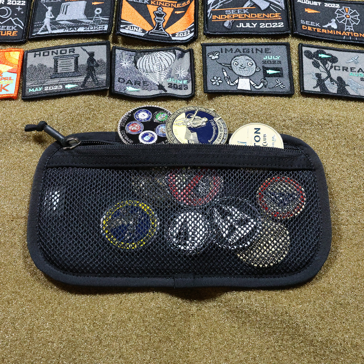 On a textured surface, a mesh pouch is filled with various collectible patches and badges. Above it, an assortment of embroidered Admin Panels from GORUCK displays different designs and words such as HONOR, IMAGINE, alongside dates from 2022—ideal for adding an adventurous flair to your travel or rucking gear.