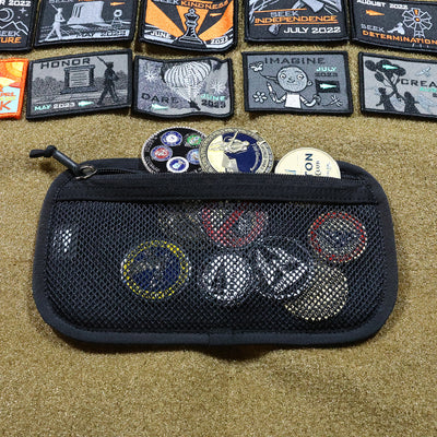 On a textured surface, a mesh pouch is filled with various collectible patches and badges. Above it, an assortment of embroidered Admin Panels from GORUCK displays different designs and words such as HONOR, IMAGINE, alongside dates from 2022—ideal for adding an adventurous flair to your travel or rucking gear.