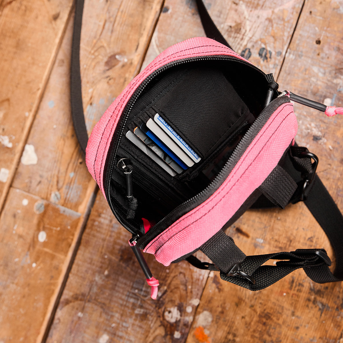 A GORUCK Admin Panels in a small pink design, perfect for everyday carry during travel, is open to reveal multiple credit cards in a black holder. It rests on a worn wooden floor with paint splatters and features a black strap and zippers.