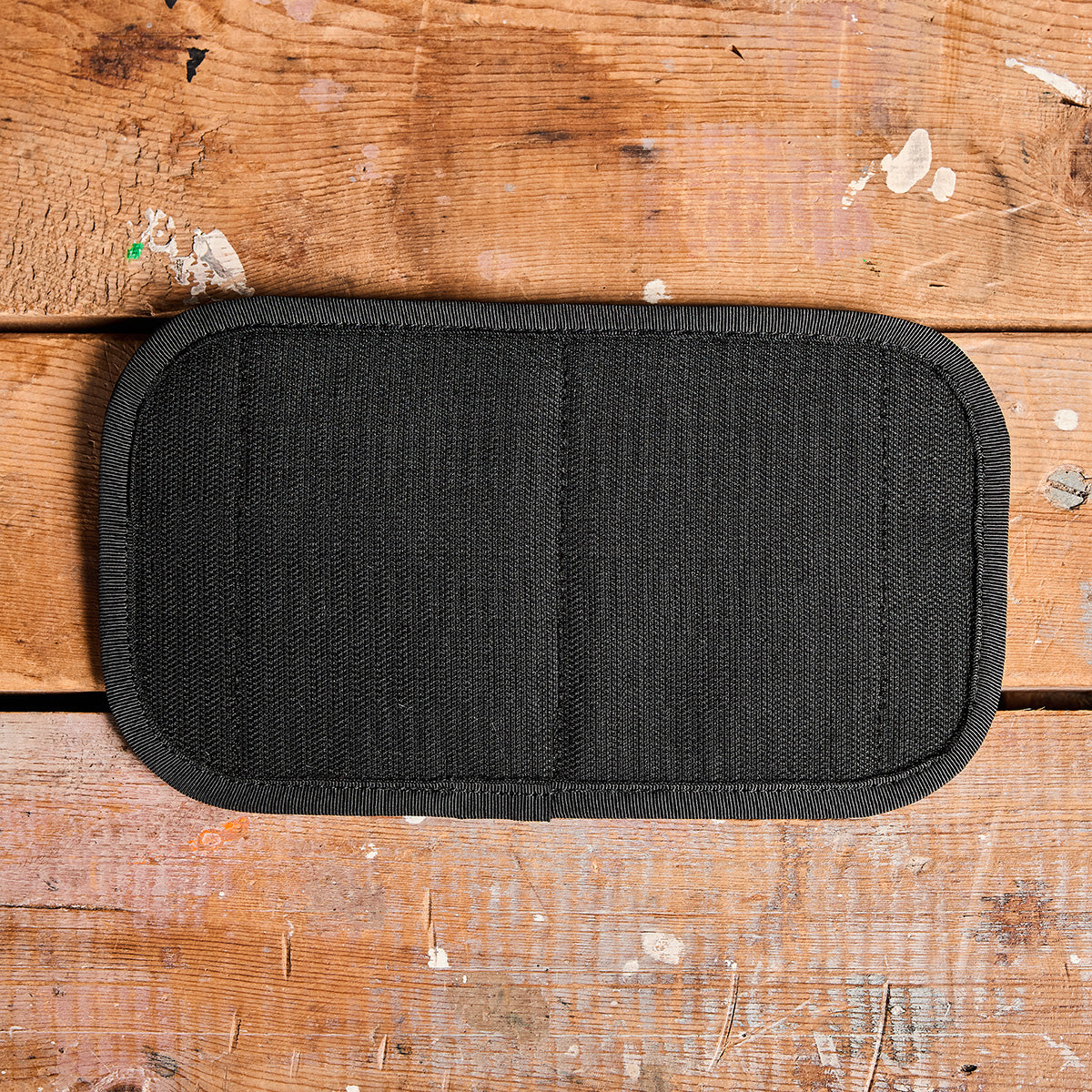 A GORUCK Admin Panel in black, featuring a rectangular fabric design with rounded edges, is showcased on a rustic wooden surface adorned with visible paint splatters and scratches, making it ideal for EDC.