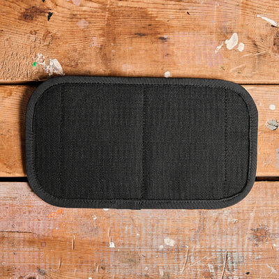 An Admin Panel by GORUCK, ideal for EDC enthusiasts or rucking adventures, lies on a worn wooden surface with scattered paint marks and scratches. The panel is black with a rectangular shape, rounded edges, and stitching down the center.