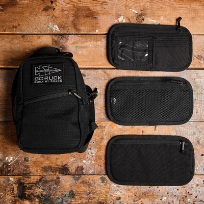 A black GORUCK Admin Panel rests on a worn wooden surface, ideal for rucking or travel. It comes with three detachable pouches—one clear, one mesh, and one solid fabric—perfect for keeping EDC items organized.