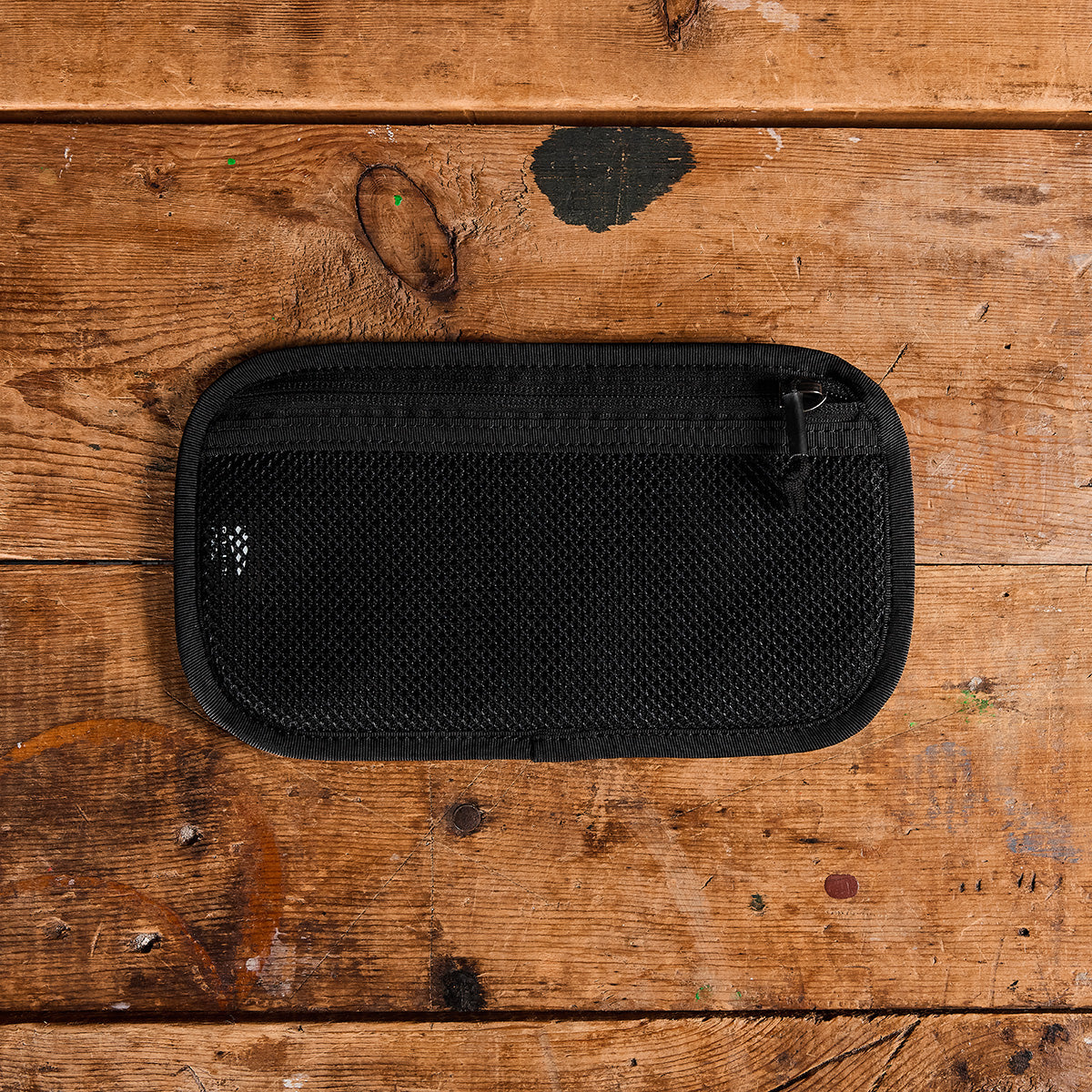 A black mesh zippered pouch from GORUCK's Admin Panels collection rests on a rustic wooden surface adorned with noticeable scratches and paint marks, ideal for EDC enthusiasts. The rectangular pouch showcases a small logo in the top left corner, seamlessly combining style and practical travel convenience.