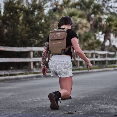 Kneeling on a road with a sturdy brown backpack, the person exudes confidence in a black shirt and GORUCK Women's Training Shorts made with ToughStretch® fabric. Surrounded by trees, they're prepared for anything, assured that their squat-proof outfit is ideal for adventure.