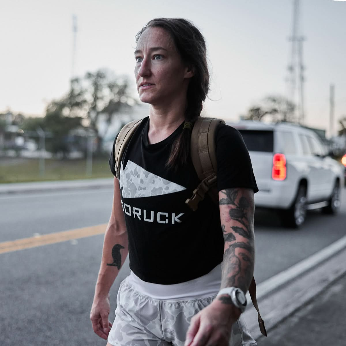 A person strides down the street, wearing a GORUCK shirt and Women's Training Shorts crafted from squat-proof ToughStretch fabric. A rugged backpack hangs over tattooed arms, embodying the spirit of adventure and resilience.