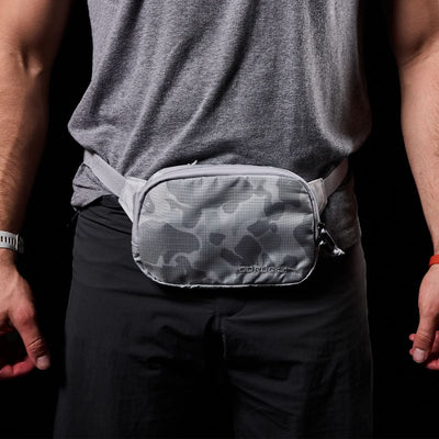 A person wearing a gray T-shirt and black pants stands against a dark background. They have the Arctic Camo Belt Bag by GORUCK, made from Ripstop ROBIC® nylon with waterproof zippers, around their waist. Their hands are relaxed by their sides.