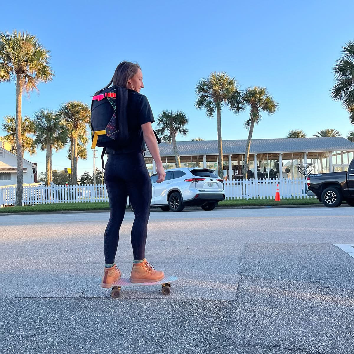 A person with a GR0 Skate Ruck - Built By SCARS from GORUCK is skateboarding down a street lined with palm trees and parked cars, their backpack secure and ready for a bombproof everyday carry. Brown boots complete their effortlessly cool look.