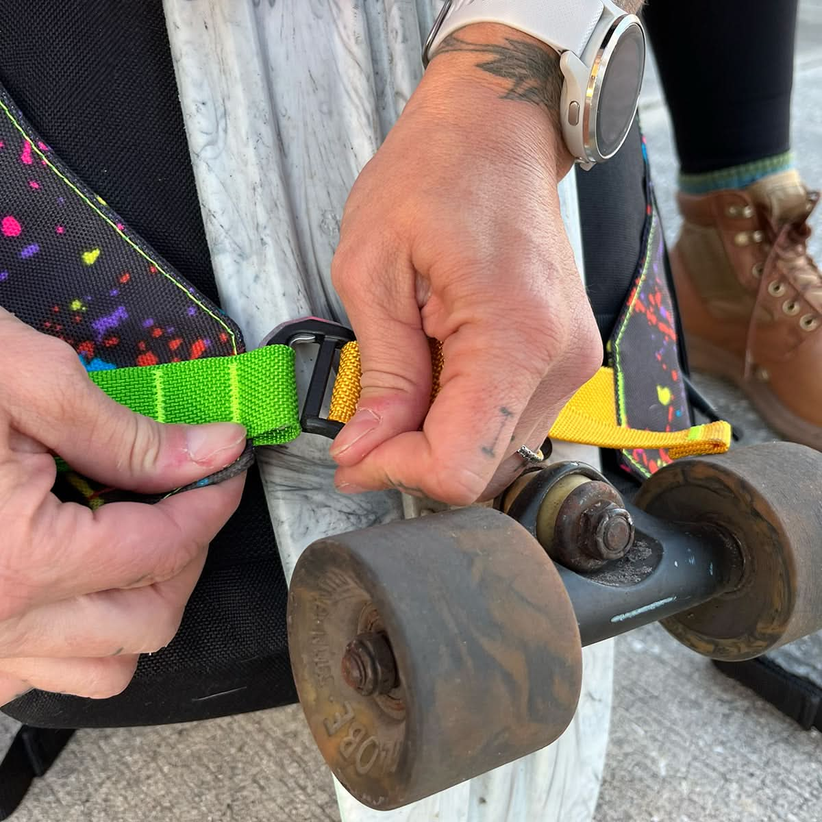 Using a vibrant strap, a skateboard is being attached to a backpack, which is part of GORUCK's GR0 Skate Ruck - Built By SCARS, designed specifically for skaters.