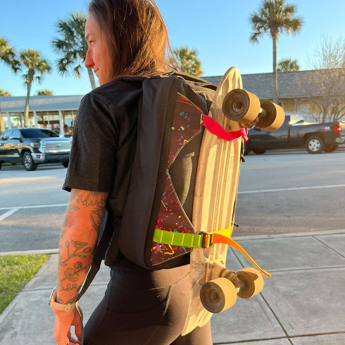 A person wearing a black shirt carries a skateboard on their GORUCK GR0 Skate Ruck - Built By SCARS backpack, customized for skaters. Palm trees and parked cars form the backdrop.