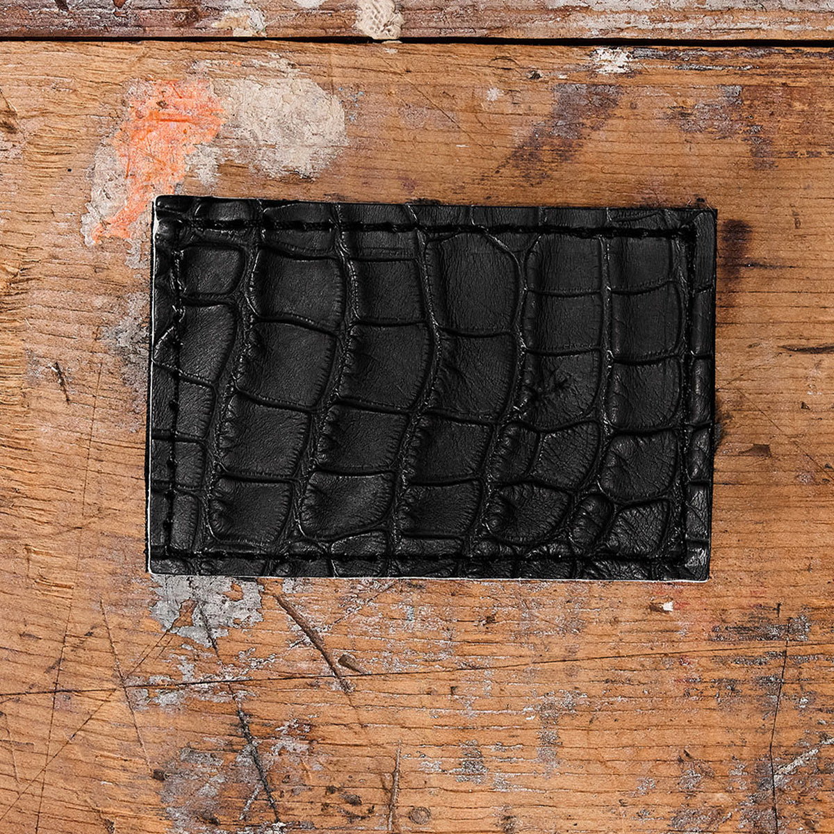 The Everglades Mystery Leather Patches wallet by GORUCK, featuring a crocodile skin pattern and textured exotic leather, rests on a weathered wooden surface marked with paint splatters and scratches.