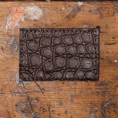 An Everglades Mystery Leather Patches card holder by GORUCK, featuring a brown exotic leather with a circular pattern, sits on a weathered wooden surface that reveals its grain and markings.