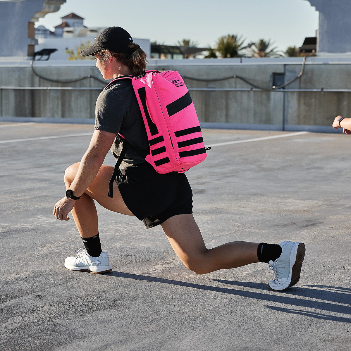 A person wearing a black cap, T-shirt, and shorts is performing a lunge on a rooftop while wearing GORUCK's Women's Ballistic Trainers in Lunar Rock + Gum with a Silver Reflective Spearhead. With a bright pink backpack made of CORDURA® Ballistic Nylon and sporting a smartwatch, they are surrounded by concrete structures with trees and rooftops extending into the distance.