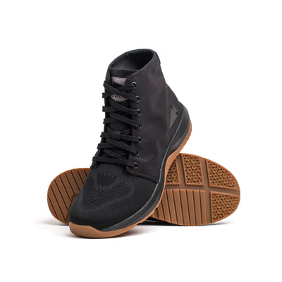 The Women's Ballistic Trainers by GORUCK, featuring a black mesh and CORDURA® Ballistic Nylon upper, are designed as mid-top sneakers with black reflective spearhead accents. These trainers boast visible laces and a gum rubber sole, with one shoe showcasing the textured sole pattern as it leans while the other stands upright—ideal for blending stability and style in functional fitness footwear.