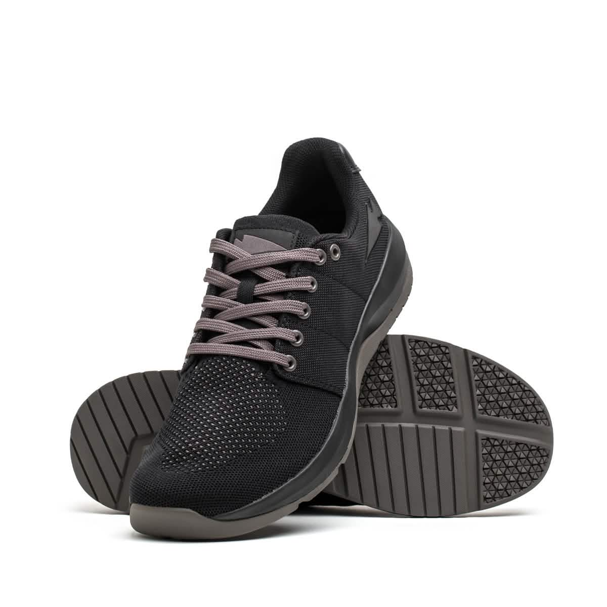 Introducing the Men's Ballistic Trainers from GORUCK in Black, Black, and Charcoal with a Black Reflective Spearhead. These athletic shoes feature textured soles tailored for functional fitness. One shoe stands upright while the other tilts slightly behind, highlighting the tread pattern. Equipped with 3X Stability technology, gray laces, and a durable mesh upper crafted from CORDURA Ballistic Nylon, these trainers are built to last.