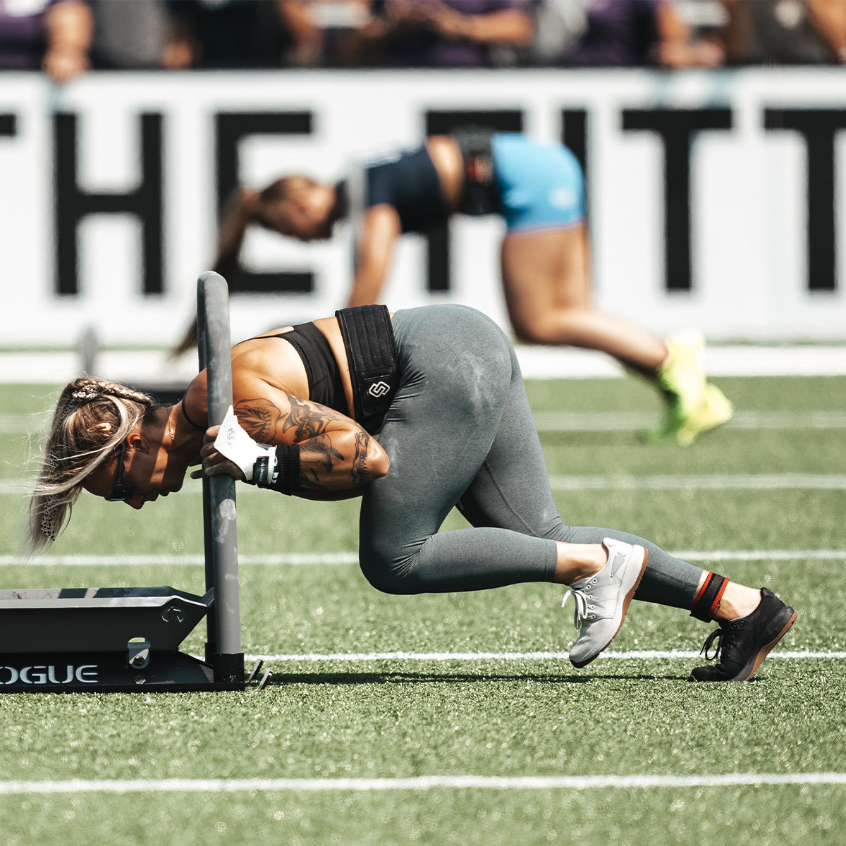 During an outdoor competition, two athletes are demonstrating their Women's Ballistic Trainers - Lunar Rock + Gum w/ Silver Reflective Spearhead by GORUCK as they push sleds. The athlete in the foreground, focused intently on her task, wears grey leggings and a black sports top. Meanwhile, the athlete in the background is sporting blue shorts while pushing a sled across the turf field.