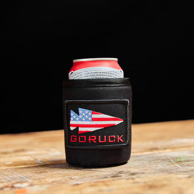 A Ballistic Beer Jacket by GORUCK, crafted from durable 1000D CORDURA in a sleek black holder with an American flag design and "GORUCK" branding, resting on a wooden surface—an ideal combination of style and resilience to maintain your beer at the perfect temperature.