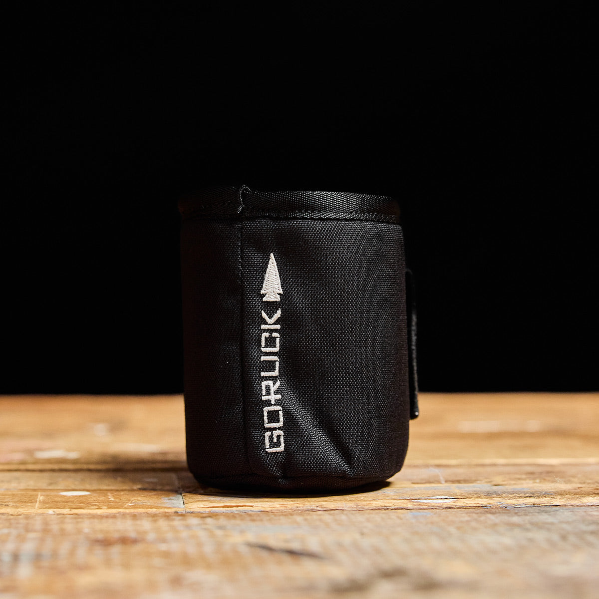 A GORUCK Ballistic Beer Jacket, made from 1000D CORDURA, sits on a wooden surface, its black color contrasting sharply with the background.