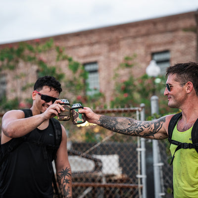 Two people with backpacks toast cold beers and smile outdoors near a brick building. Their Ballistic Beer Jackets from GORUCK, crafted from durable 1000D CORDURA, seem ready for any adventure that comes their way.